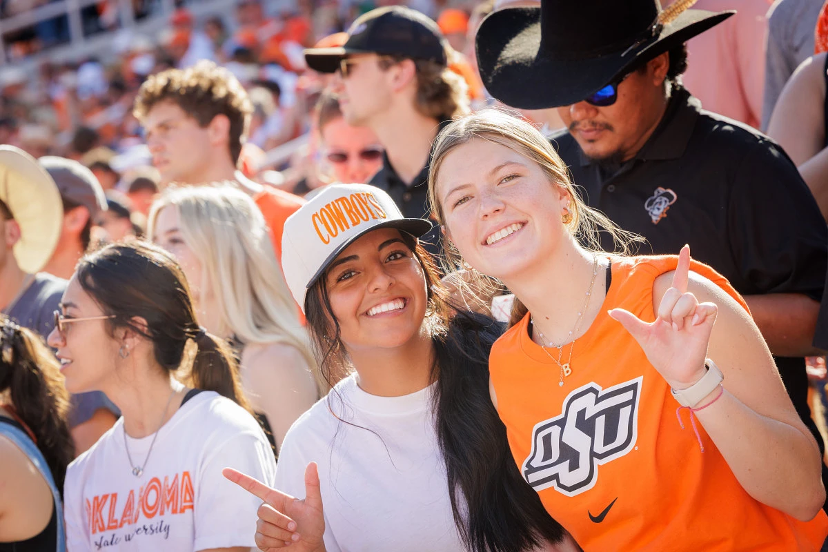 Two smiling friends dressed in Oklahoma State University attire pose together at a crowded outdoor event.